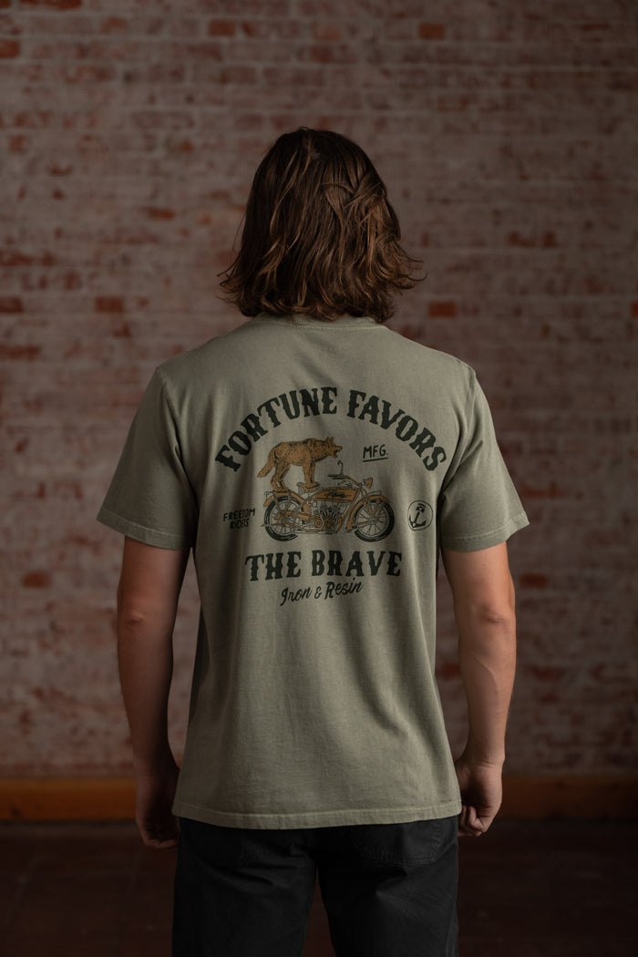 Man in olive shirt with "Fortune Favors the Brave" text and motorcycle graphic, standing against a brick wall.