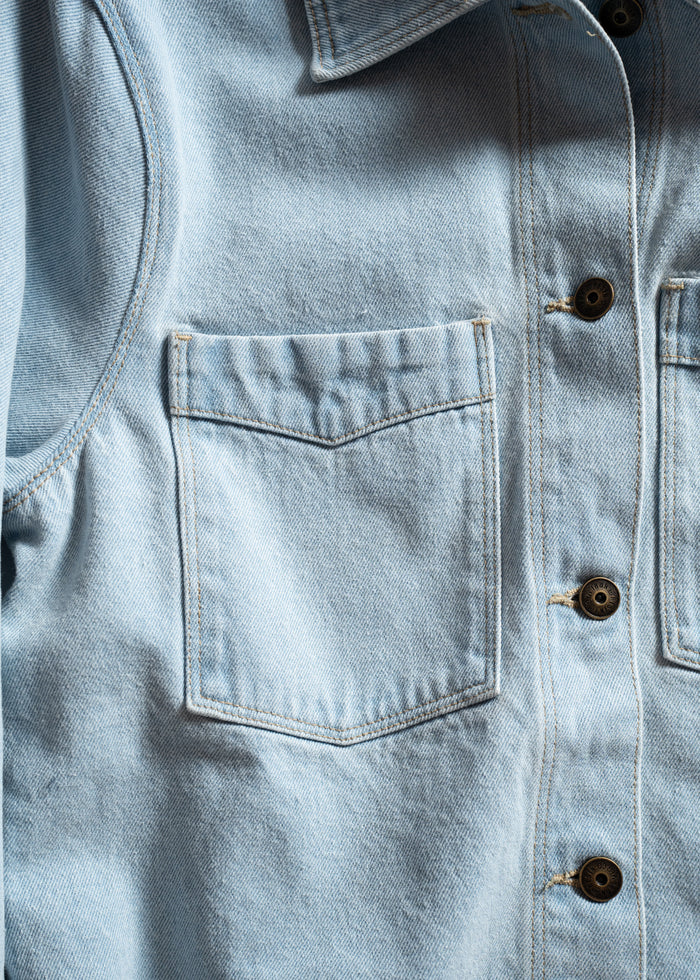 Close-up of a light blue denim shirt with a front pocket and buttons, highlighting the texture and stitching details.