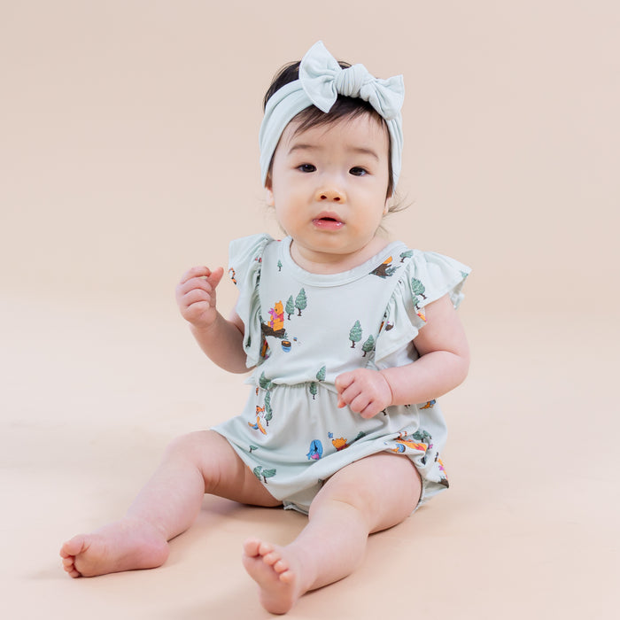 Young toddler sitting on the floor wearing the Bubble Romper in Winnie the Pooh Hundred Acre Wood paired with a light green bow