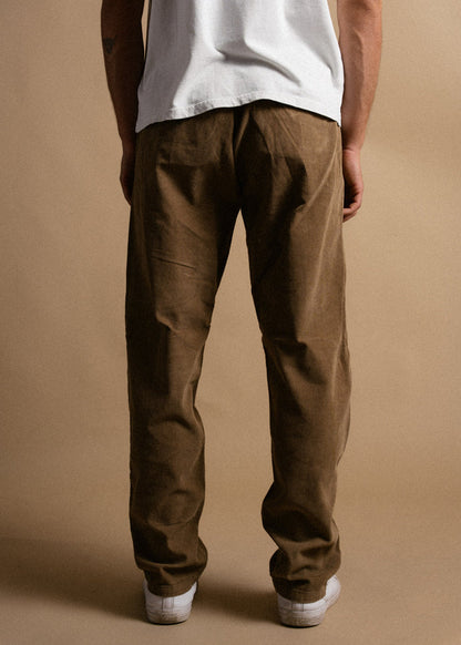 Man in brown corduroy pants and white sneakers stands on a beige background, wearing a white t-shirt.
