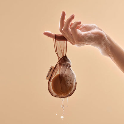 Hand holding the Kitsch Chocolate Body Wash Bar Bag in the shower with water dripping from the mesh bag