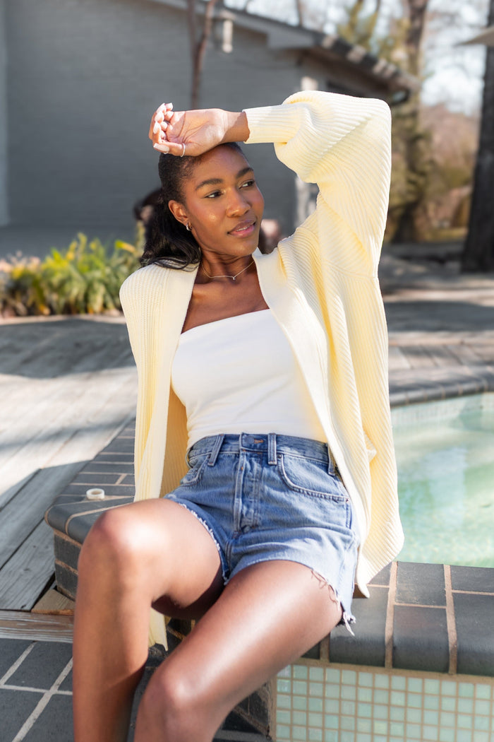 Female model sitting pool side with her hand on her forehead wearing the Chunky Knit Women's Oversized Cardigan in Chamomile with a white shirt and medium wash jean shorts