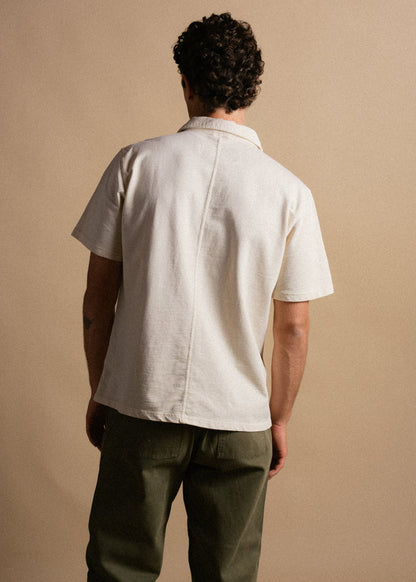 Man in a white short-sleeve shirt and green pants stands with back to camera against a beige background, showcasing casual style.