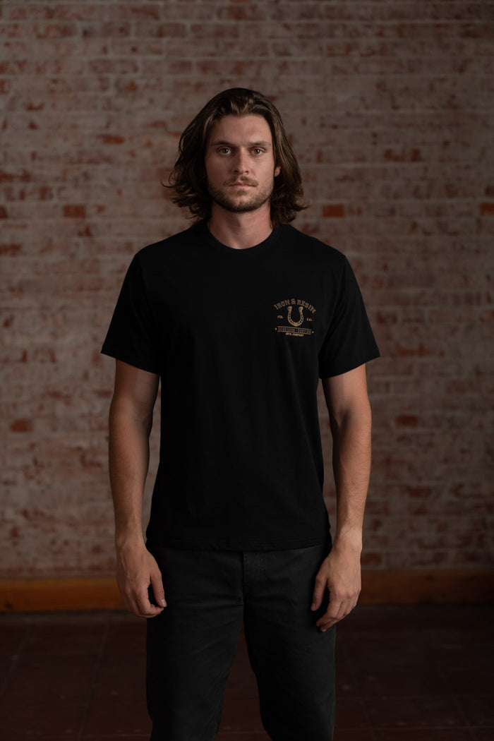 Man in a black t-shirt stands against a brick wall, looking forward. The shirt has a small logo on the left chest.