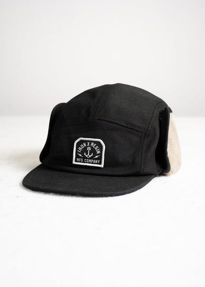 Black cap with ear flaps, featuring "Iron & Resin MFG Company" logo on front, displayed on a white surface.