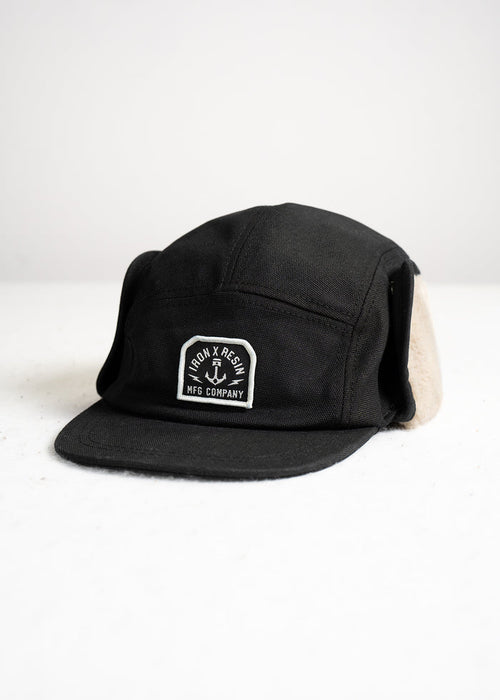 Black cap with ear flaps, featuring "Iron & Resin MFG Company" logo on front, displayed on a white surface.