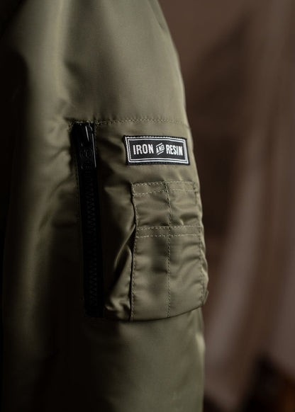 Close-up of an olive green jacket sleeve with a black zipper and "Iron and Resin" patch, highlighting its rugged style.