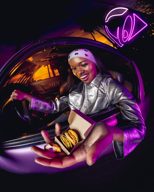 Person in a car holding Miniverse Taco Bell tacos with a neon Taco Bell logo sign in the background