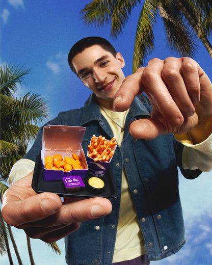 Person holding a box of Miniverse Taco Bell chicken nuggets and nacho fries with palm trees and blue sky in the background