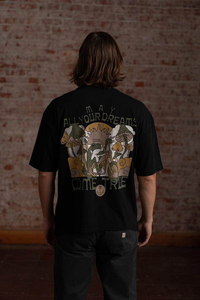 Man in black shirt with "May All Your Dreams Come True" text and mushroom design, standing against a brick wall background.