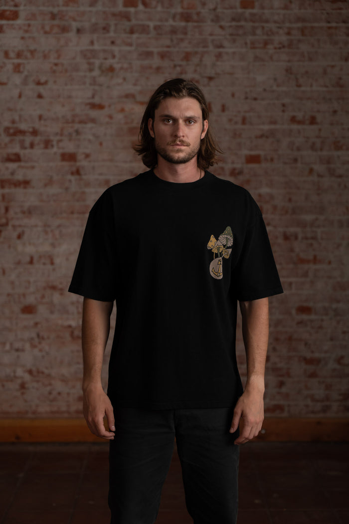 Man in black t-shirt with mushroom design stands against a brick wall, looking directly at the camera with a serious expression.