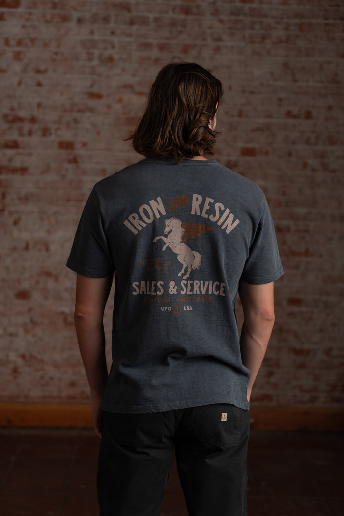 Man in a blue "Iron and Resin" shirt with a winged horse design stands against a brick wall, facing away.
