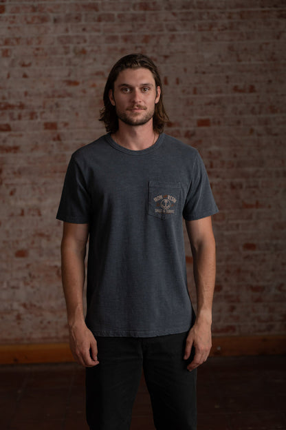 Man in a dark t-shirt stands against a rustic brick wall, looking directly at the camera with a calm expression.