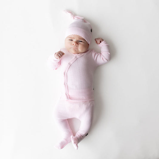 Newborn laying on a white surface wearing the Pointelle Take Me Home Set with Cap in Sakura with the picot trim visible