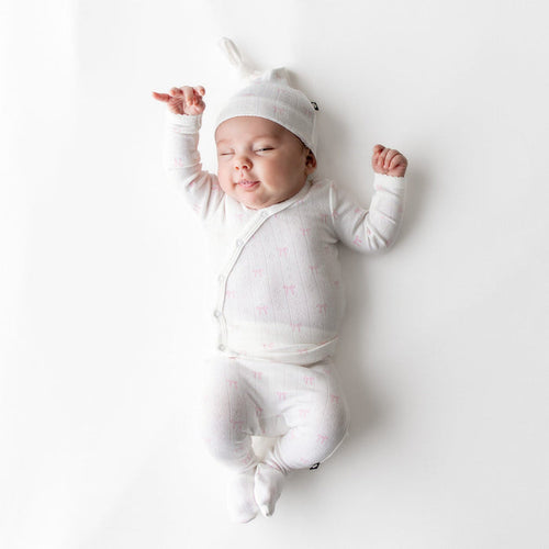 Newborn baby in a white outfit and hat with a pink bow pattern lying on a white background