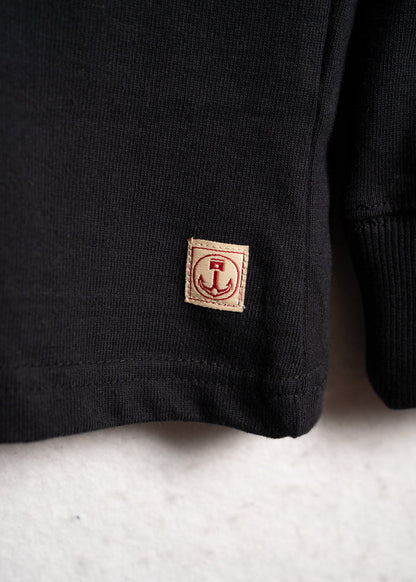 Close-up of a black sweater's hem with a small beige patch featuring a red anchor design, set against a light background.