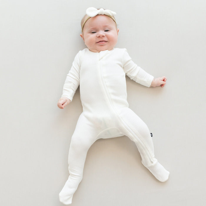 Infant laying on off-white surface wearing Baby Ribbed Zipper Footie Ribbed Zipper Footie in Cloud with a Kyte Baby Bow in Cloud