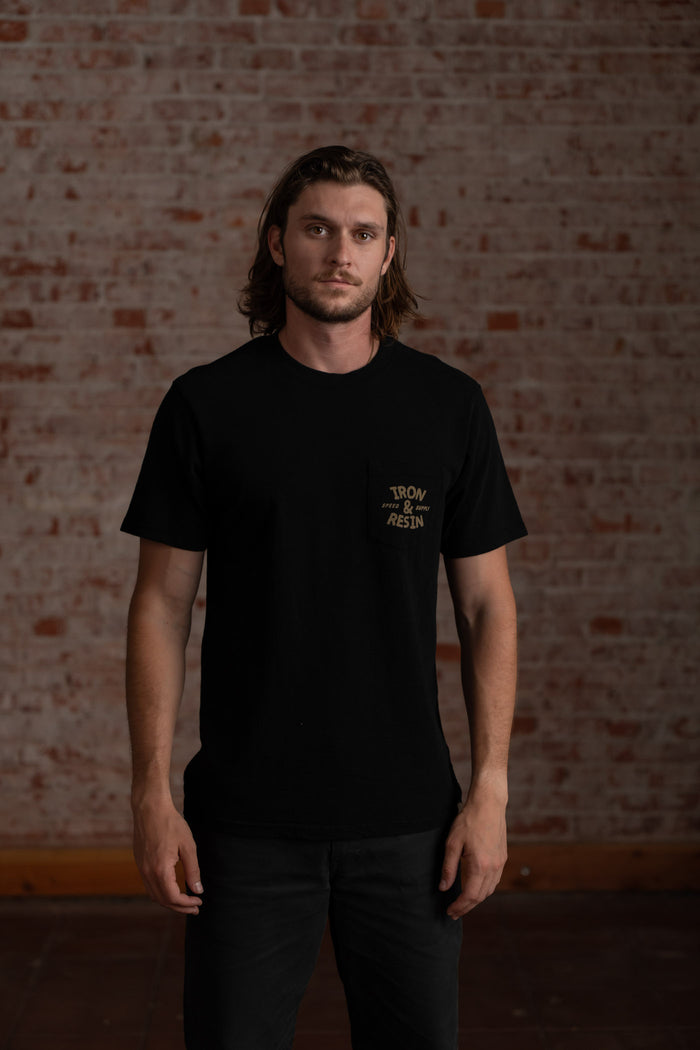Man in black "Iron & Resin" shirt stands against a brick wall, exuding a casual, confident vibe.