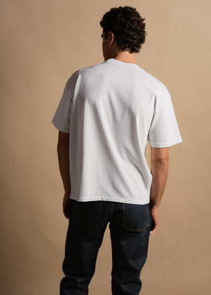 Man in white t-shirt and dark jeans stands with back to camera against beige background, showcasing casual fashion style.