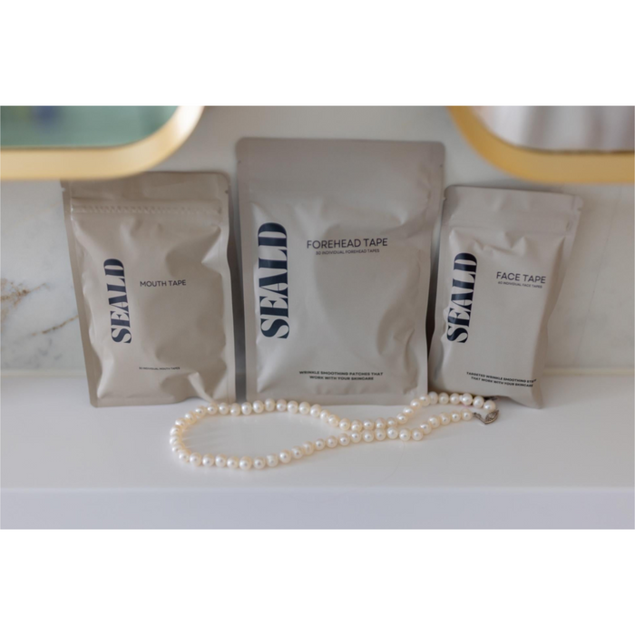 Three matte pouches of SEALD brand face and mouth tape arranged on a white surface, with a pearl necklace placed in front. The center pouch is labeled FOREHEAD TAPE, with the side pouches labeled MOUTH TAPE and FACE TAPE.