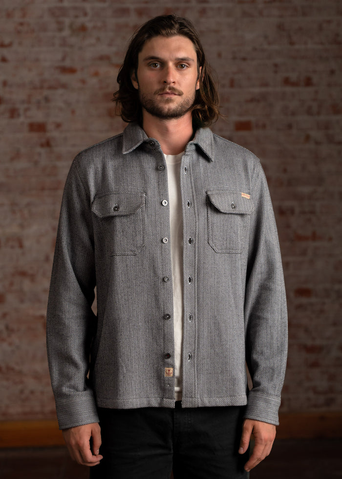 Man in a gray button-up shirt with a subtle pattern stands against a brick wall, looking directly at the camera.
