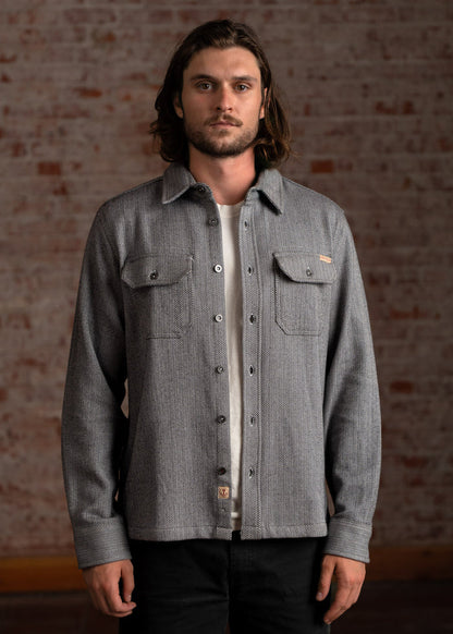 Man in a gray button-up shirt with a subtle pattern stands against a brick wall, looking directly at the camera.
