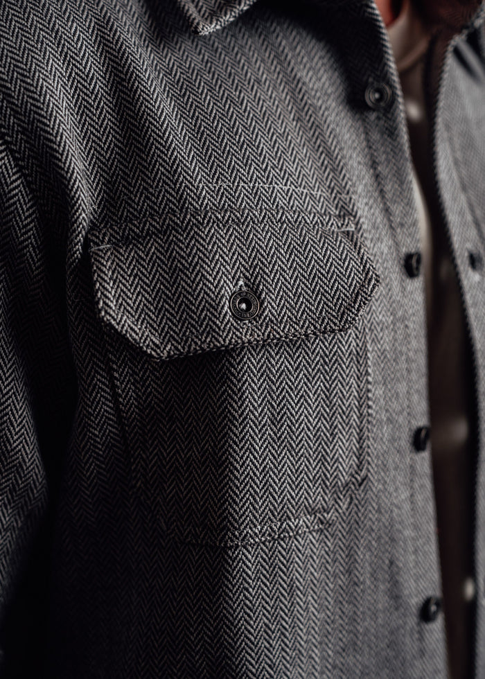 Close-up of a herringbone-patterned gray jacket with a buttoned chest pocket, highlighting texture and fabric detail.