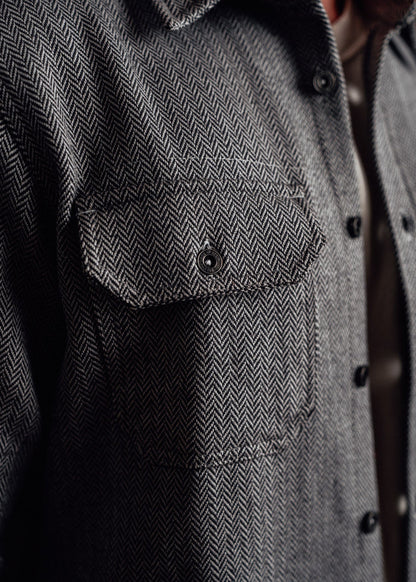 Close-up of a herringbone-patterned gray jacket with a buttoned chest pocket, highlighting texture and fabric detail.