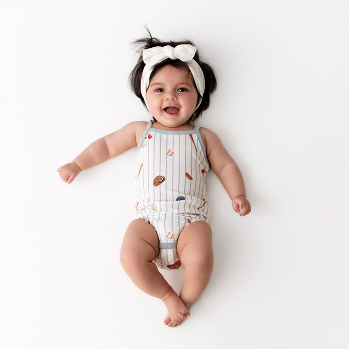 Infant girl laying on a white surface wearing the Spaghetti Strap Leotard in Home Run with a white knotted bow headband
