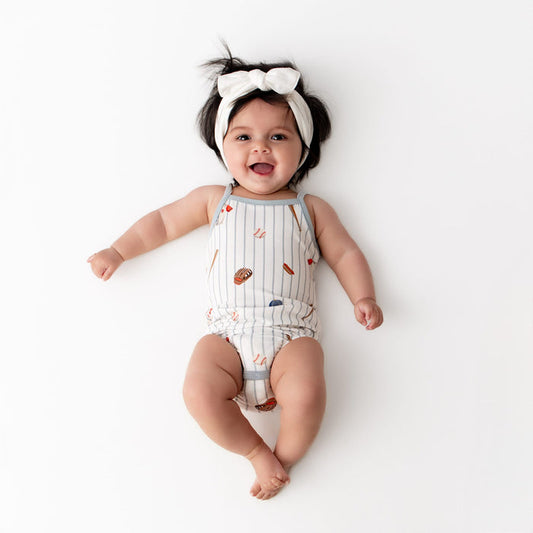 Infant girl laying on a white surface wearing the Spaghetti Strap Leotard in Home Run with a white knotted bow headband