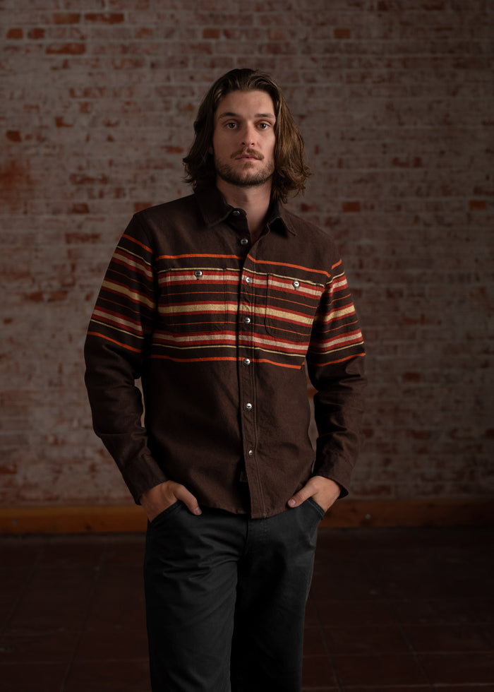 Man in a brown striped shirt stands confidently against a rustic brick wall, hands in pockets, exuding a relaxed, stylish vibe.