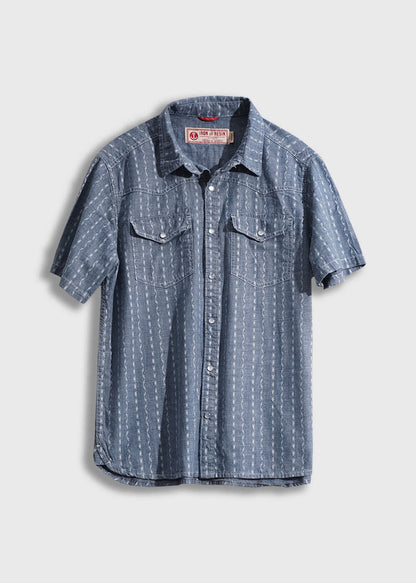 Blue short-sleeve denim shirt with white patterns, button front, and two chest pockets, hanging against a plain background.