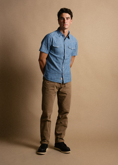 Man in a blue short-sleeve shirt and khaki pants stands against a beige background, hands in pockets, looking at the camera.