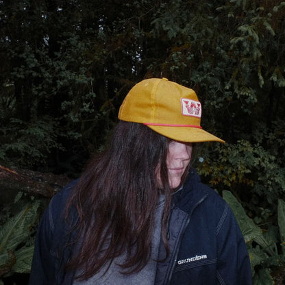Person wearing a yellow cap with a logo, standing in front of green foliage.