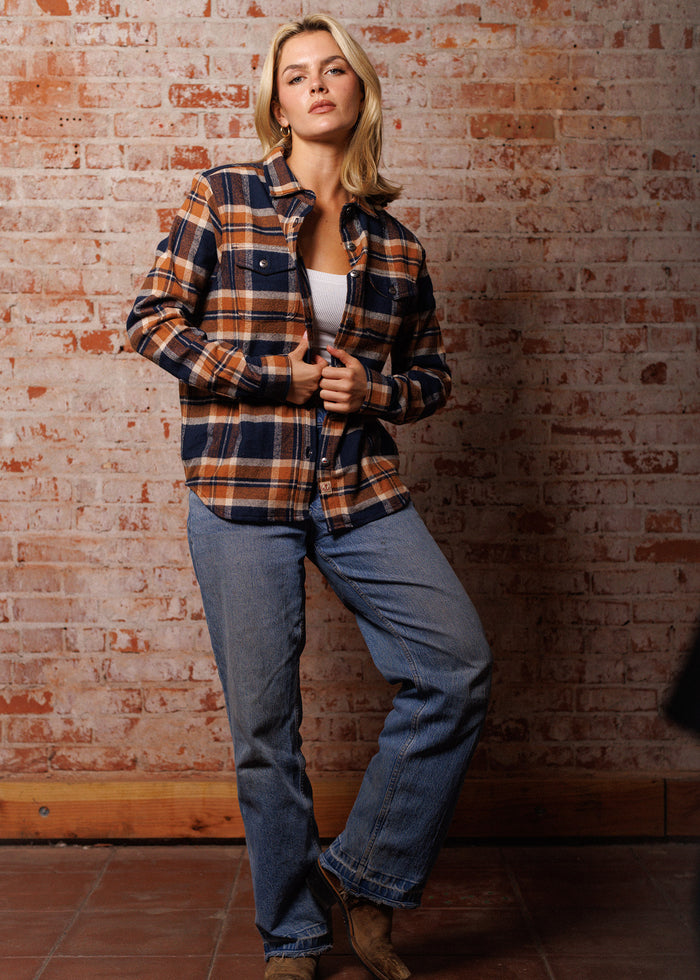 Woman in plaid shirt and jeans stands confidently against a rustic brick wall, exuding casual style and relaxed elegance.