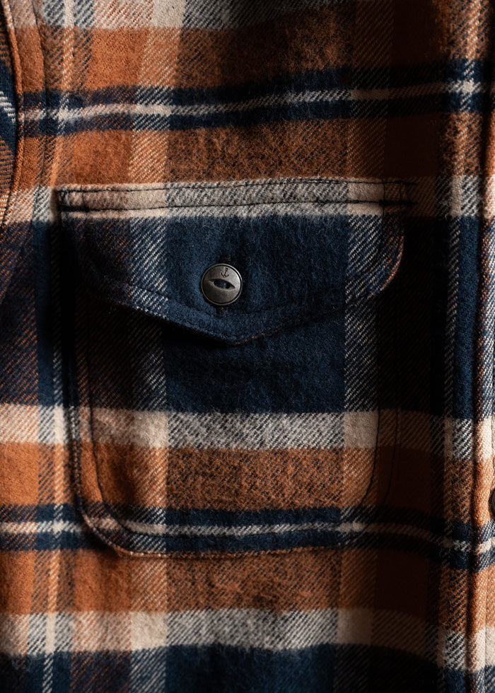 Close-up of a plaid shirt pocket with blue, orange, and white checks, featuring a button closure.