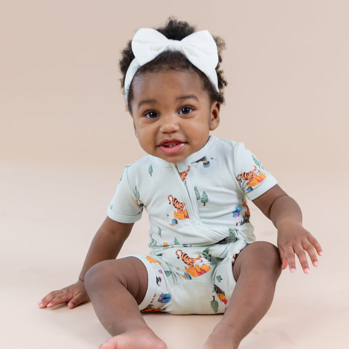 Toddler sitting on the floor wearing the Zipper Shortall Romper in Winnie the Pooh Hundred Acre Wood with a white bow