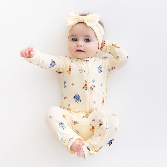 Young toddler girl stretching wearing the Zippered Romper in Winnie the Pooh Celebration with light yellow bow
